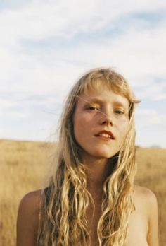 a woman with long blonde hair standing in the middle of an open field looking at the camera