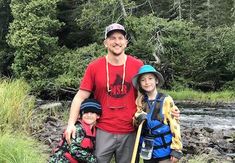 a man and two children standing next to each other near a river with trees in the background