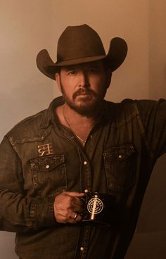 a man wearing a cowboy hat and holding a camera in his right hand while standing next to a wall