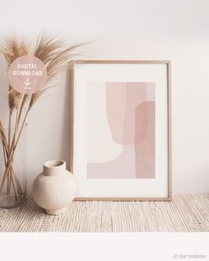 a white vase sitting next to a painting on top of a wooden table near a plant