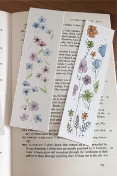 three bookmarks with flowers painted on them sitting next to an open book and some pages