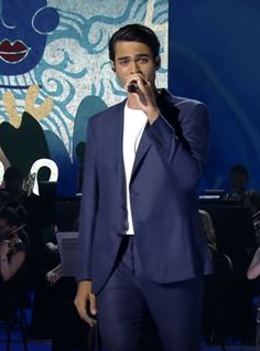 a man in a blue suit singing into a microphone with an audience behind him watching