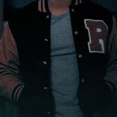 a man wearing a baseball jacket and glasses standing in front of a wall with his hands in his pockets