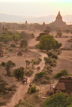 people riding horses down a dirt road in the middle of an area with trees and buildings