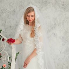 a woman in a white wedding dress holding a red rose on her finger and wearing a veil over her head