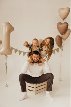 a man sitting on top of a wooden crate with two women and a baby in front of balloons