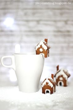 three small gingerbread houses sitting next to a white coffee cup and saucer in the snow