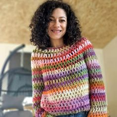 a woman wearing a multicolored crocheted sweater and jeans stands in front of a wall