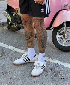 a man with tattoos on his legs standing next to a pink scooter and moped