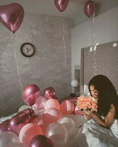 a woman sitting on a bed with balloons floating in the air above her and holding a bouquet of flowers