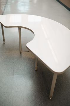 a curved white table sitting on top of a gray floor next to a wall with windows