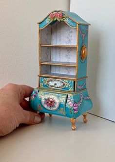 a hand is holding a miniature blue and gold cabinet