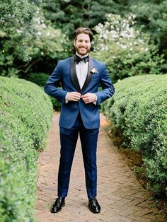 a man in a tuxedo standing on a brick path surrounded by green bushes