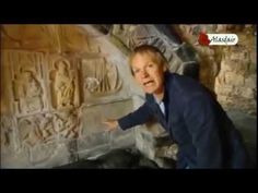 the man is pointing at an ancient wall with carvings on it and his hands are out