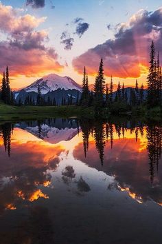 the sun is setting over a mountain with trees in front of it and clouds reflecting in the water