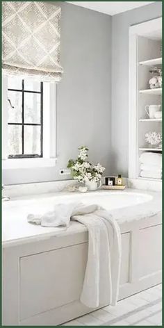 a white bath tub sitting under a window next to a shelf filled with towels and flowers