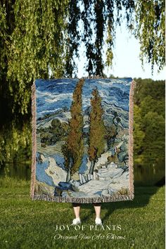 a woman standing in the grass holding up a blanket that has trees on it and mountains behind her