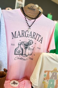 shirts and necklaces are on display in a store window, including one for margarita