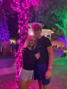 a man and woman standing next to each other in front of trees with lights on them