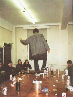 a group of people sitting around a wooden table with cans on the floor and one person standing up