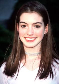 a woman with long brown hair wearing a white shirt and diamond choker smiling at the camera