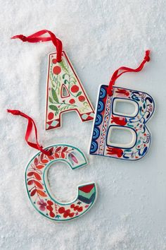 three christmas ornaments are hanging on the snow covered ground, one is decorated with flowers and the other has a letter shaped ornament