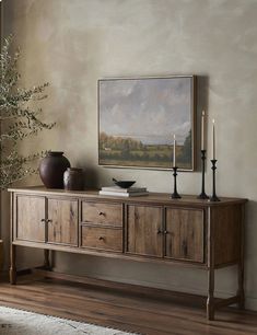 a large painting hangs on the wall above a wooden table with candles and vases