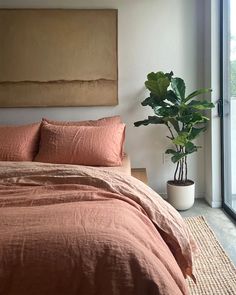 a bed with an orange comforter next to a large painting and a potted plant