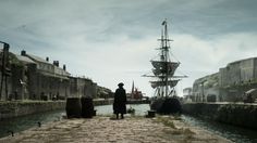 a man standing on the side of a river next to a tall ship