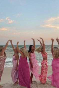 four women in pink dresses on the beach with their arms in the air and one holding her hands up