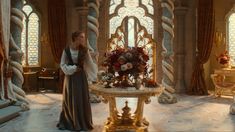 a woman standing next to a table with flowers on it in a room filled with windows