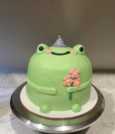 a green cake decorated with flowers and a smiling frog on it's side, sitting on a silver platter