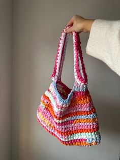 a hand holding a multicolored crocheted bag in front of a white wall