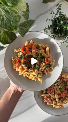 two plates of pasta with tomatoes and spinach on them, one being held up by someone's hand