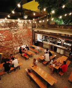 people are sitting at tables in an open area with lights strung from the brick wall