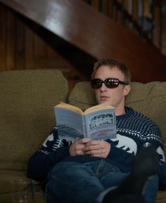 a young man sitting on a couch reading a book with sunglasses on his face and looking at the camera