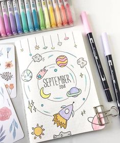 an open notebook next to markers and pens on a white surface with space themed stickers