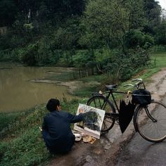 a man sitting on the side of a road next to a bike and holding an easel