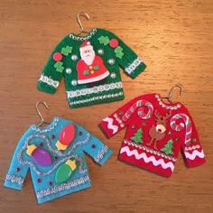 three christmas sweater ornament hanging from hooks on a wooden table with other ornaments