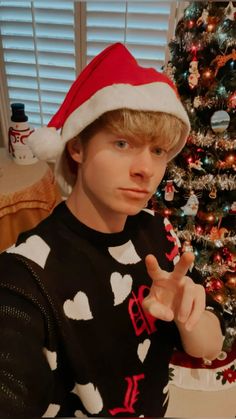 a young man wearing a christmas sweater and santa hat making the peace sign in front of a decorated christmas tree