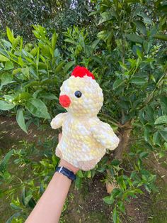 a hand is holding a stuffed chicken in front of some bushes and trees with green leaves