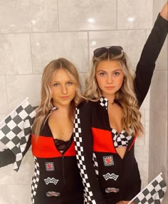 two women dressed in racing outfits posing for the camera with their arms up and one holding a checkered flag