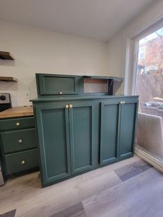 an empty kitchen with green cabinets and wood flooring in front of a large window