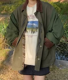 a woman standing in front of a pond wearing a green jacket and white shirt with an animal print on it