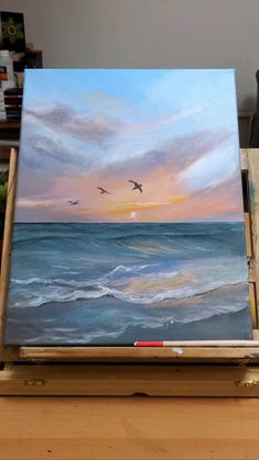 an easel with a painting on it and two birds flying over the ocean at sunset