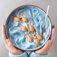 two hands holding a bowl filled with blue liquid and gold fish on top of it