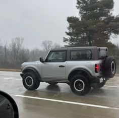 a silver four door suv driving down a road on a rainy day with trees in the background