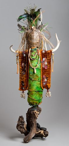 a sculpture made out of various items on top of a tree stump with feathers and beads hanging from it's sides