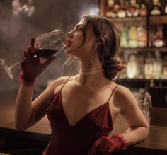 a woman in a red dress drinking from a wine glass while sitting at a bar