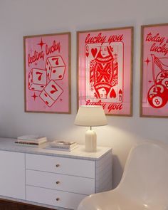 two framed pictures hang on the wall above a white dresser with a lamp and chair